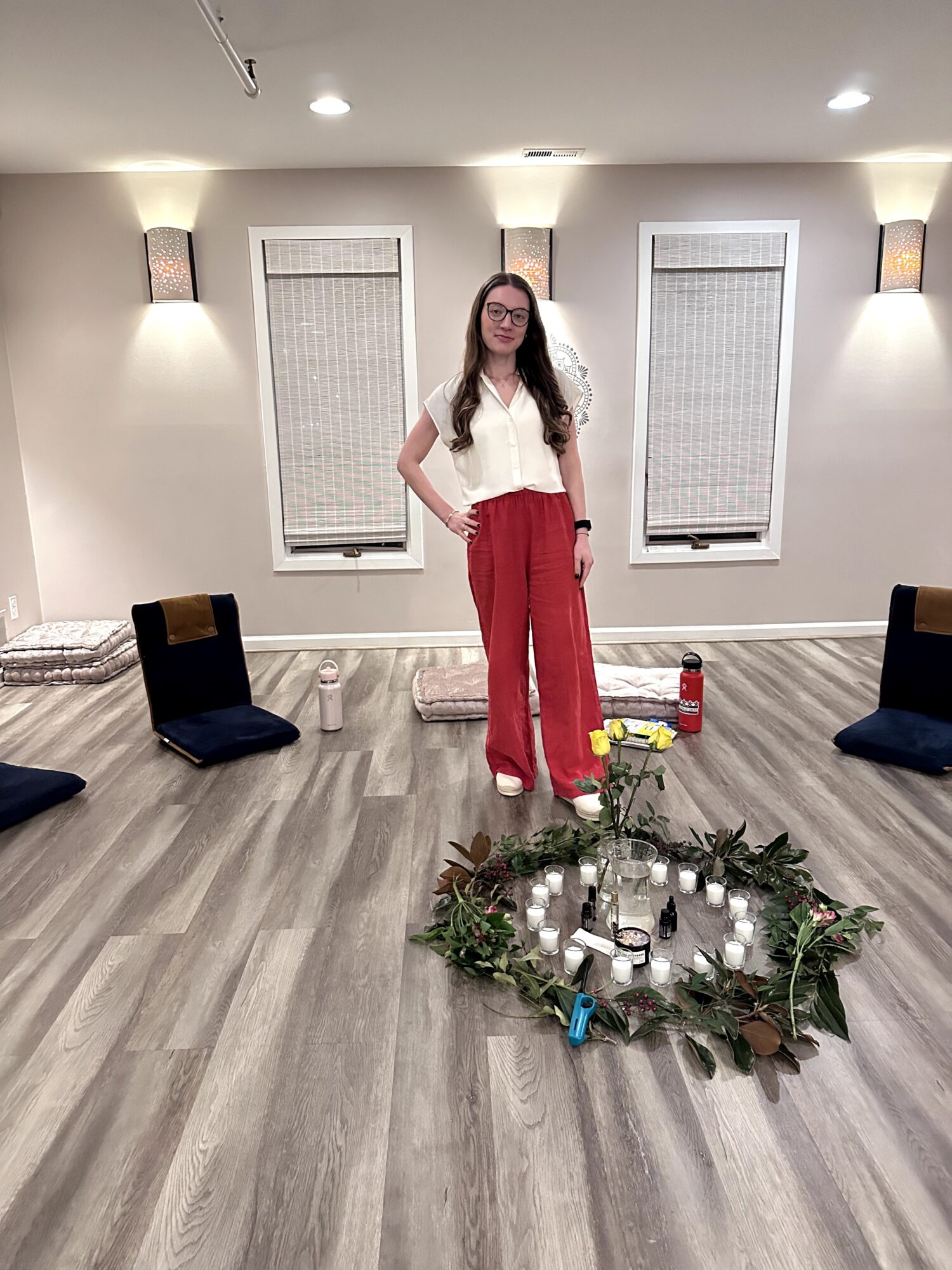 Woman standing in a decorated room with candles and greenery, wearing a white top and red pants, with windows and wall lights behind her.