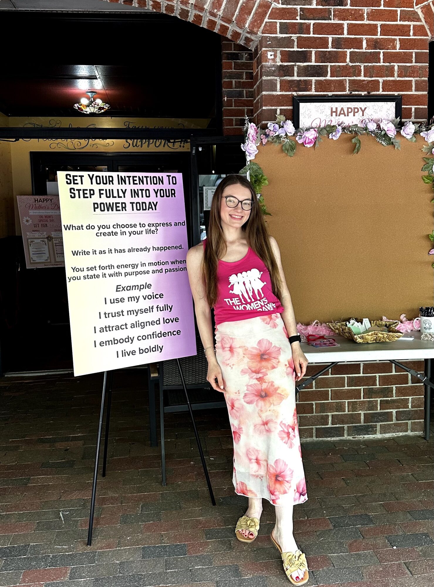 Young woman with glasses and long hair, wearing a pink top and floral skirt, standing next to a whiteboard and decorated backdrop.