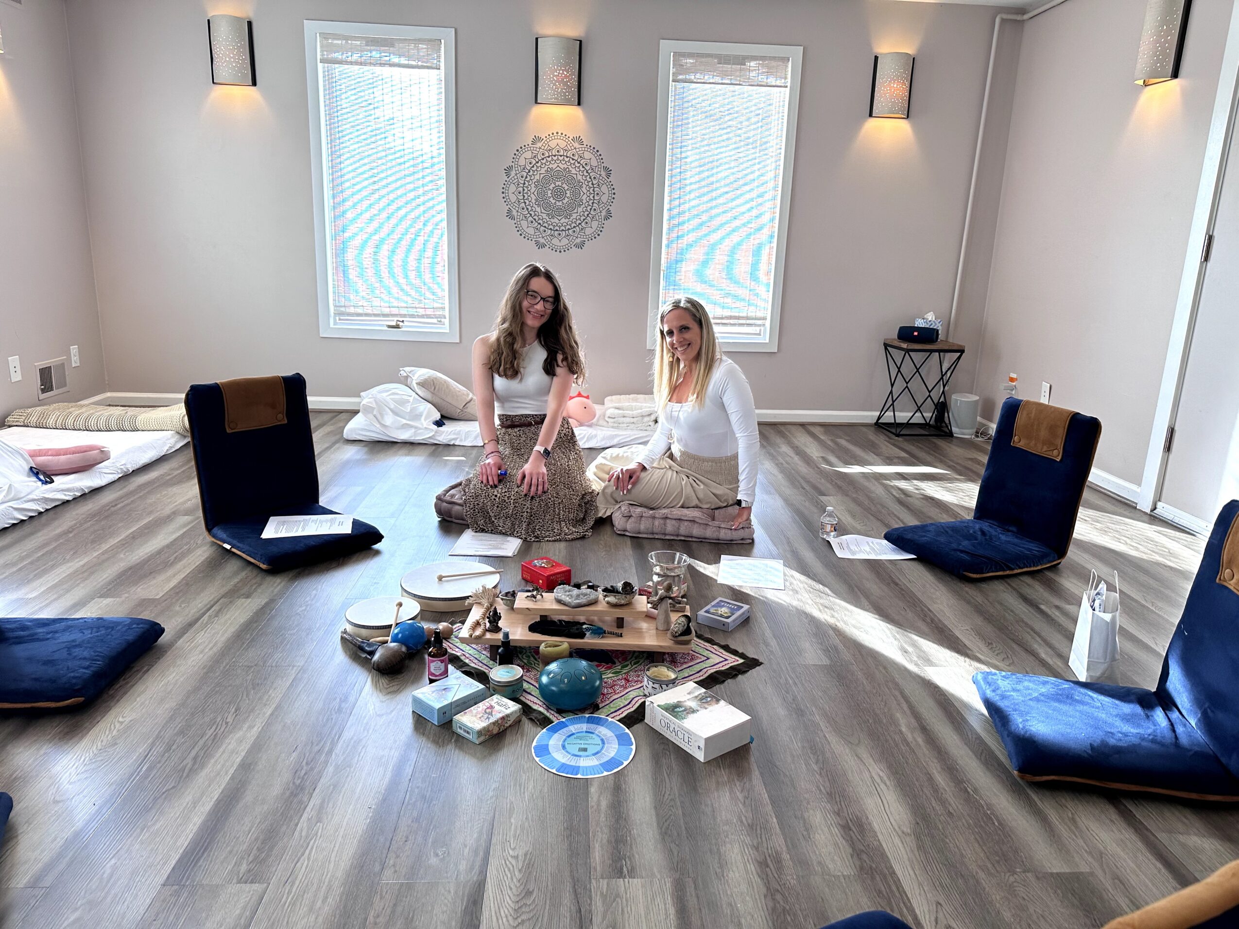 Two women sit on the floor in a bright room with three windows, surrounded by cushions and various objects on the floor.