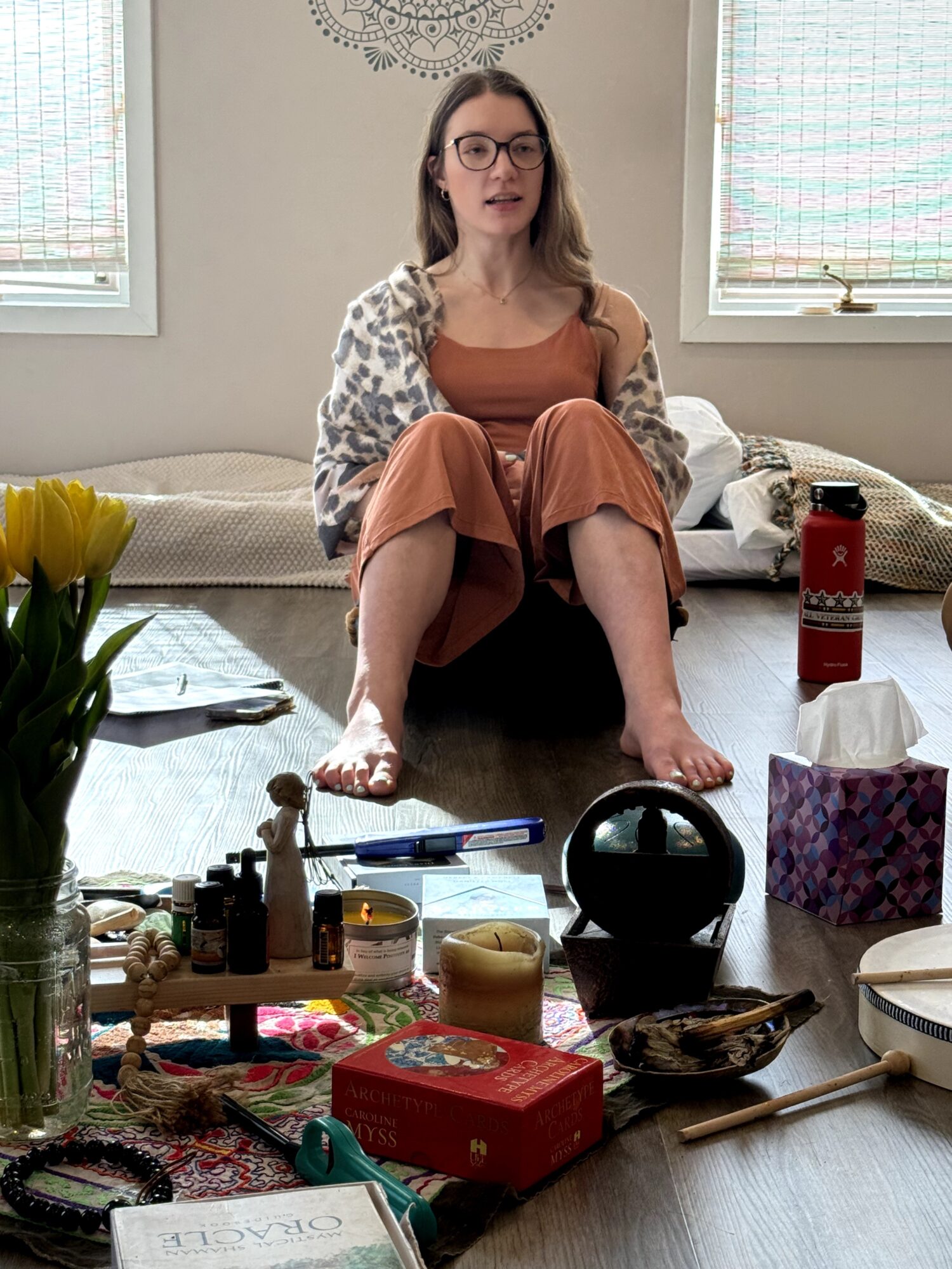 Woman sitting on a table with legs crossed, wearing glasses and a brown dress, in a room with windows and various objects on the table.