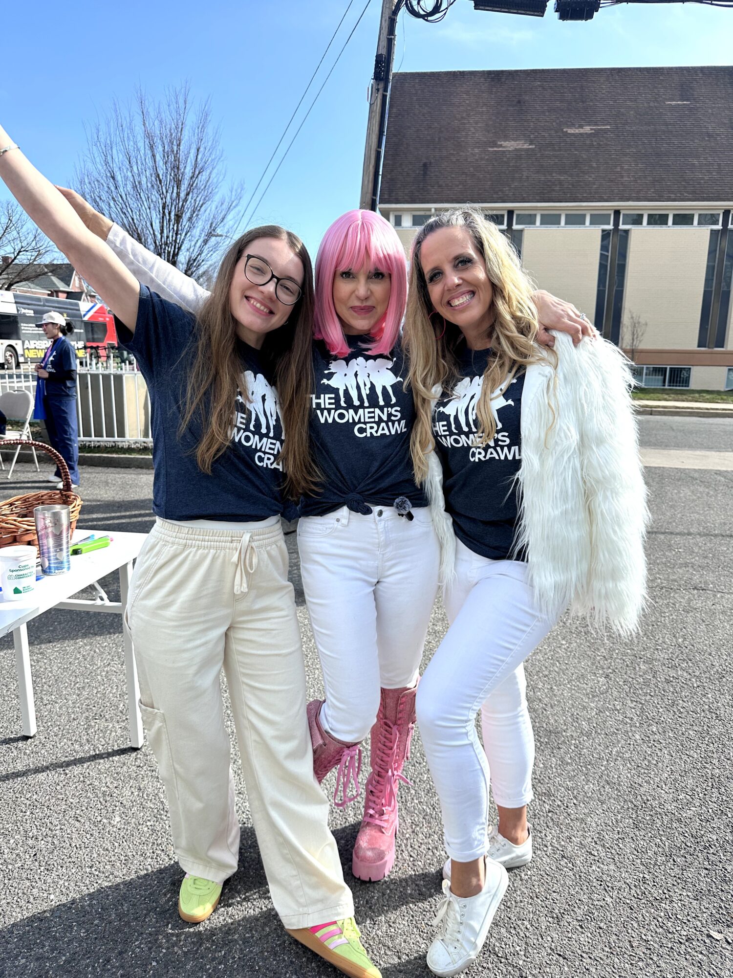 Three women standing outdoors, smiling, with one woman in pink hair and pink boots, wearing matching dark shirts.