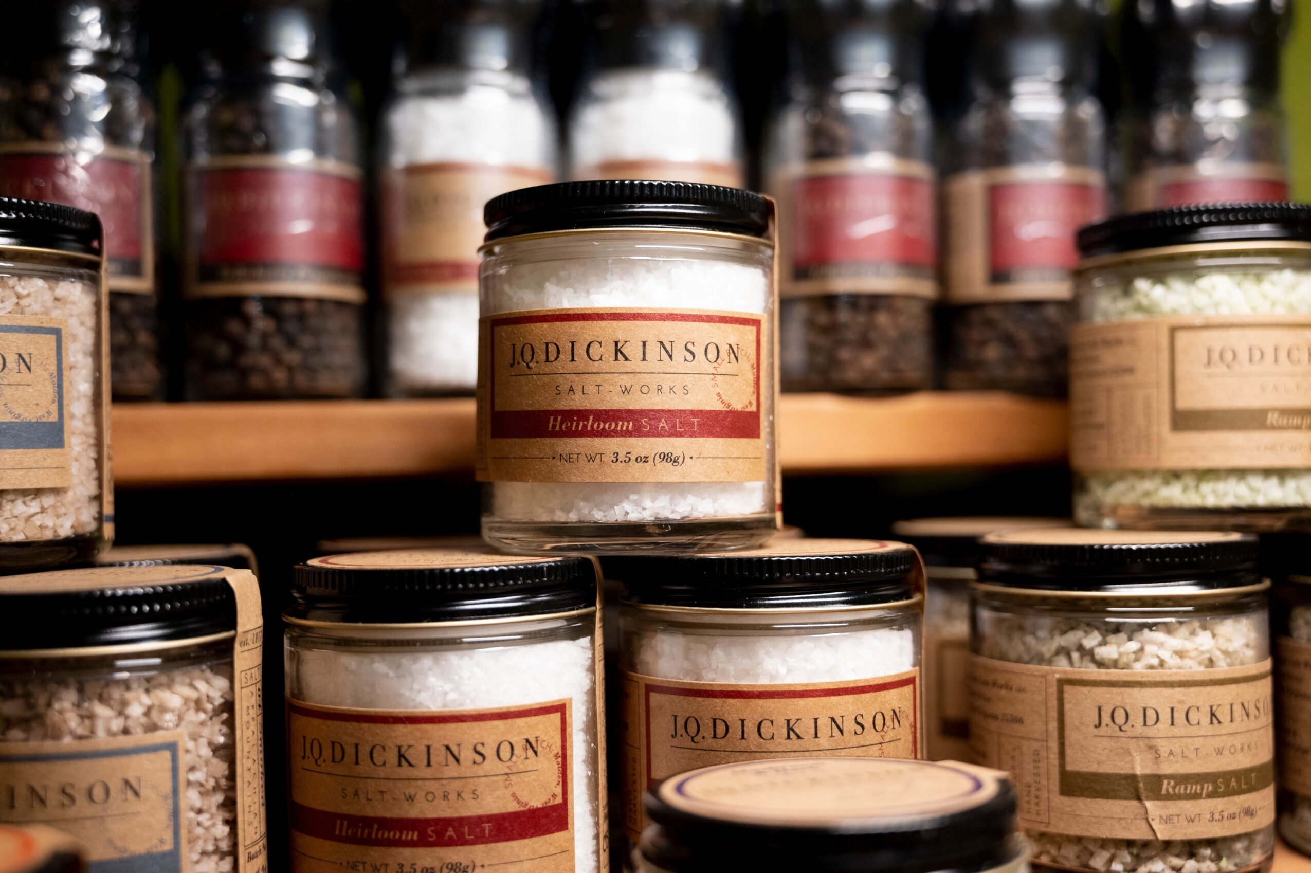 Multiple jars of spices on a shelf, with labels and black lids, some jars stacked behind others.