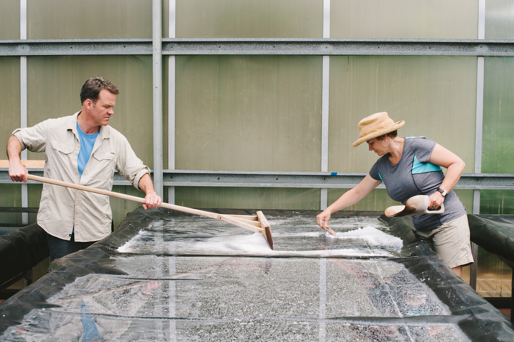 Two people clean a large outdoor surface with a long-handled tool, one wearing a hat and the other with short hair.