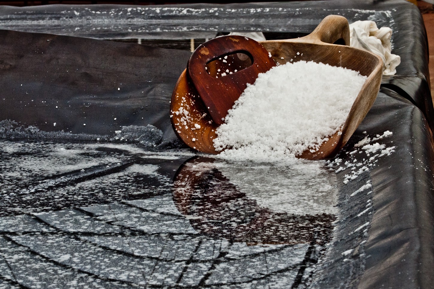 Wooden scoop filled with white salt spilling onto a black surface, with a cloth nearby.