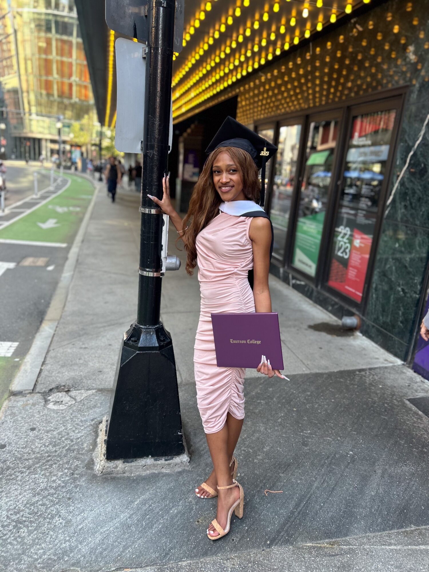 Woman in pink dress holding a diploma standing on city sidewalk near a pole and storefronts.