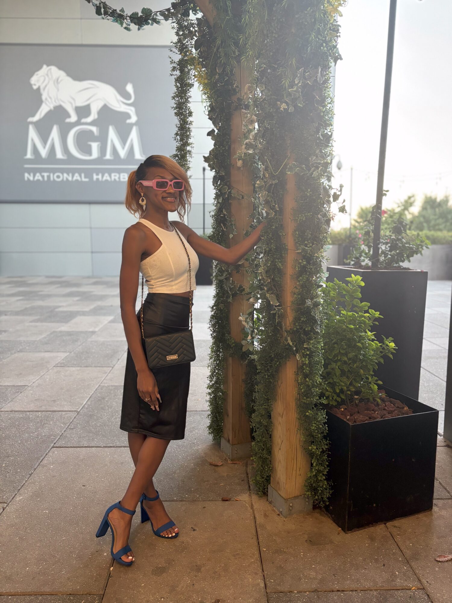 Woman in sunglasses and high heels standing next to a vertical plant display outside MGM National Harbor, holding a purse.