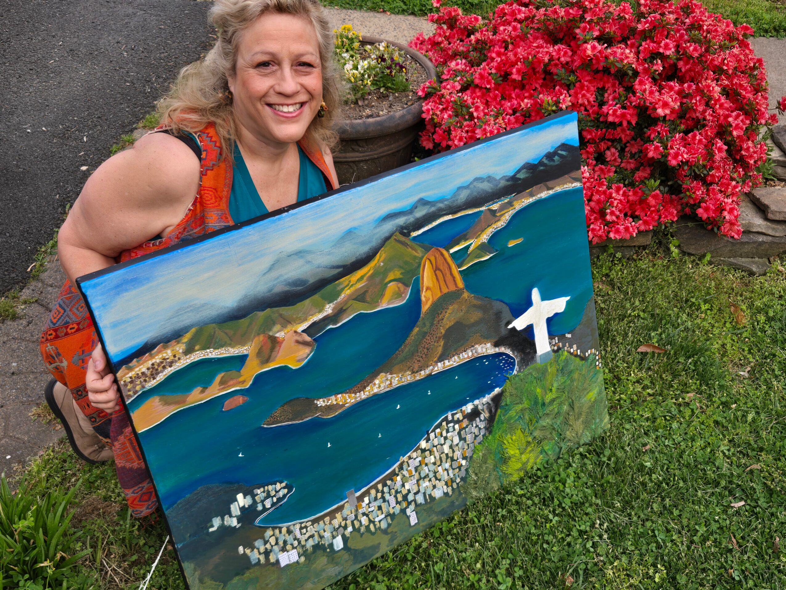 Woman smiling outdoors holding a colorful landscape painting with water, hills, and a white cross, next to pink flowers.