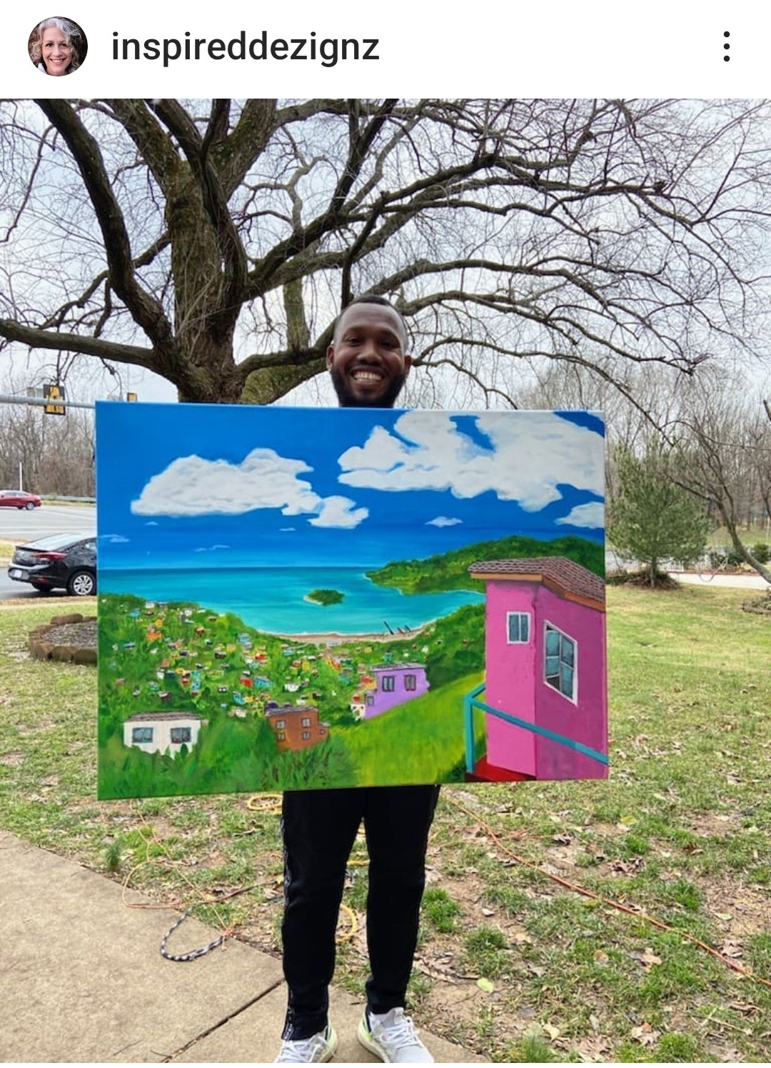 Person holding a colorful landscape painting outdoors with trees and a park in the background.