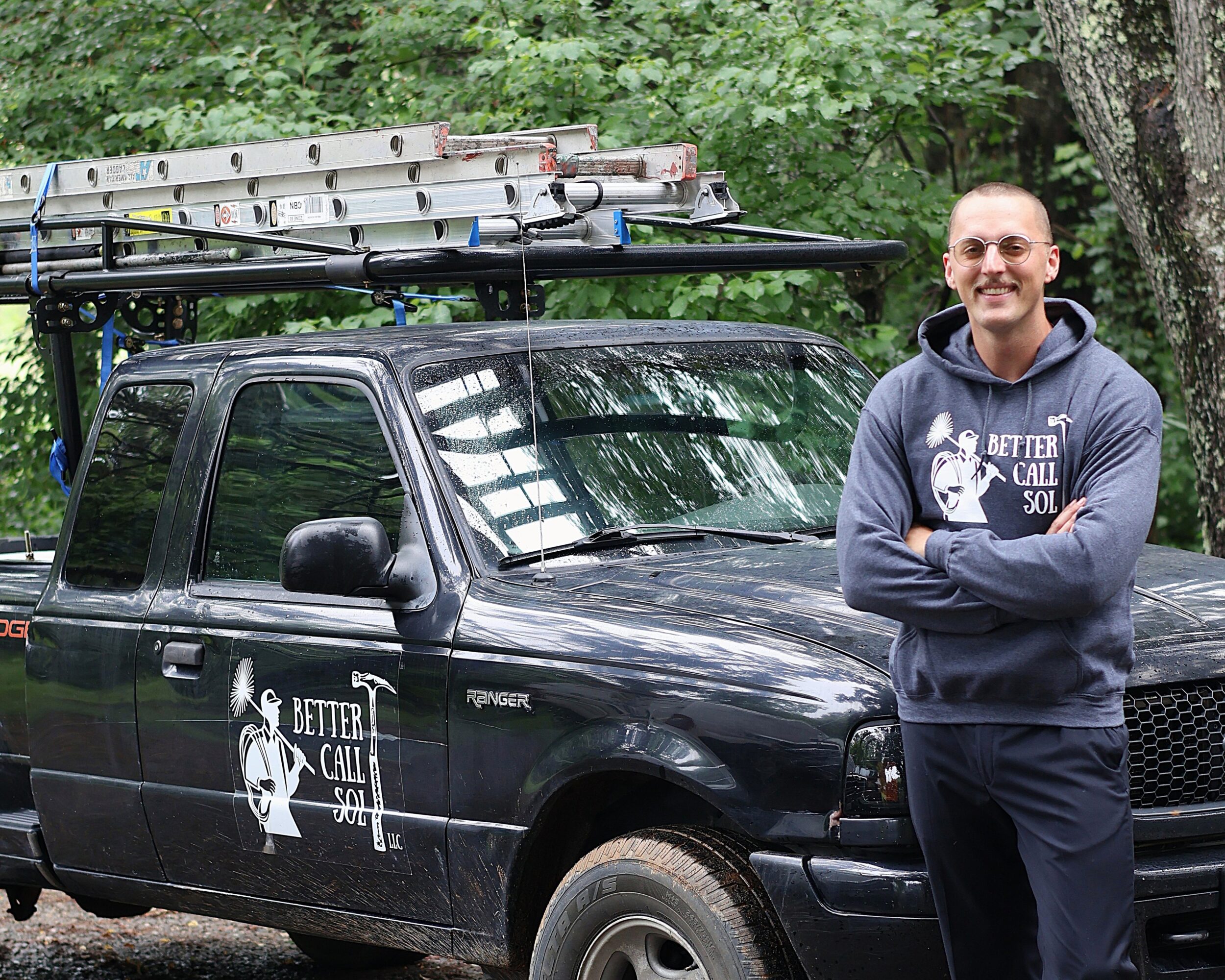Man standing next to a black pickup truck with a ladder on top, outdoors with trees in the background.