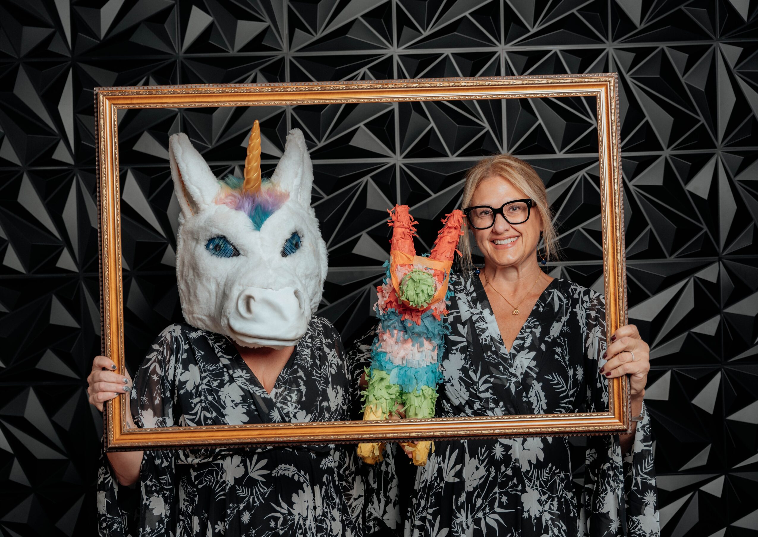 Two women, one in a unicorn mask, holding a rainbow-colored llama plush, inside a wooden frame against a black geometric background.