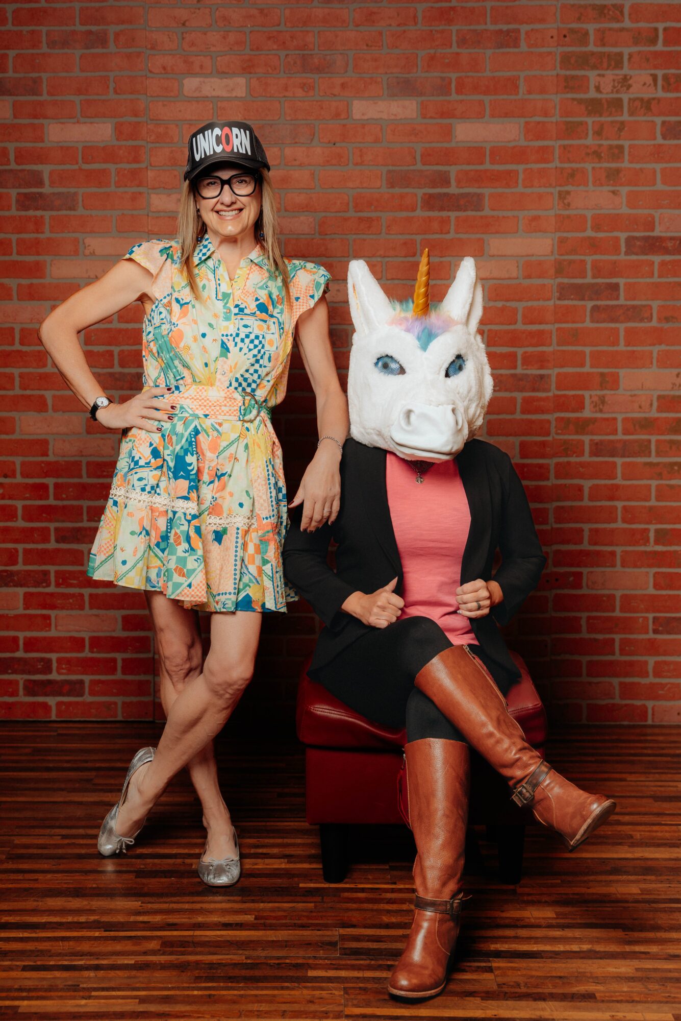 Woman in colorful dress and glasses standing next to a person in a unicorn mask sitting on a red chair, brick wall background.