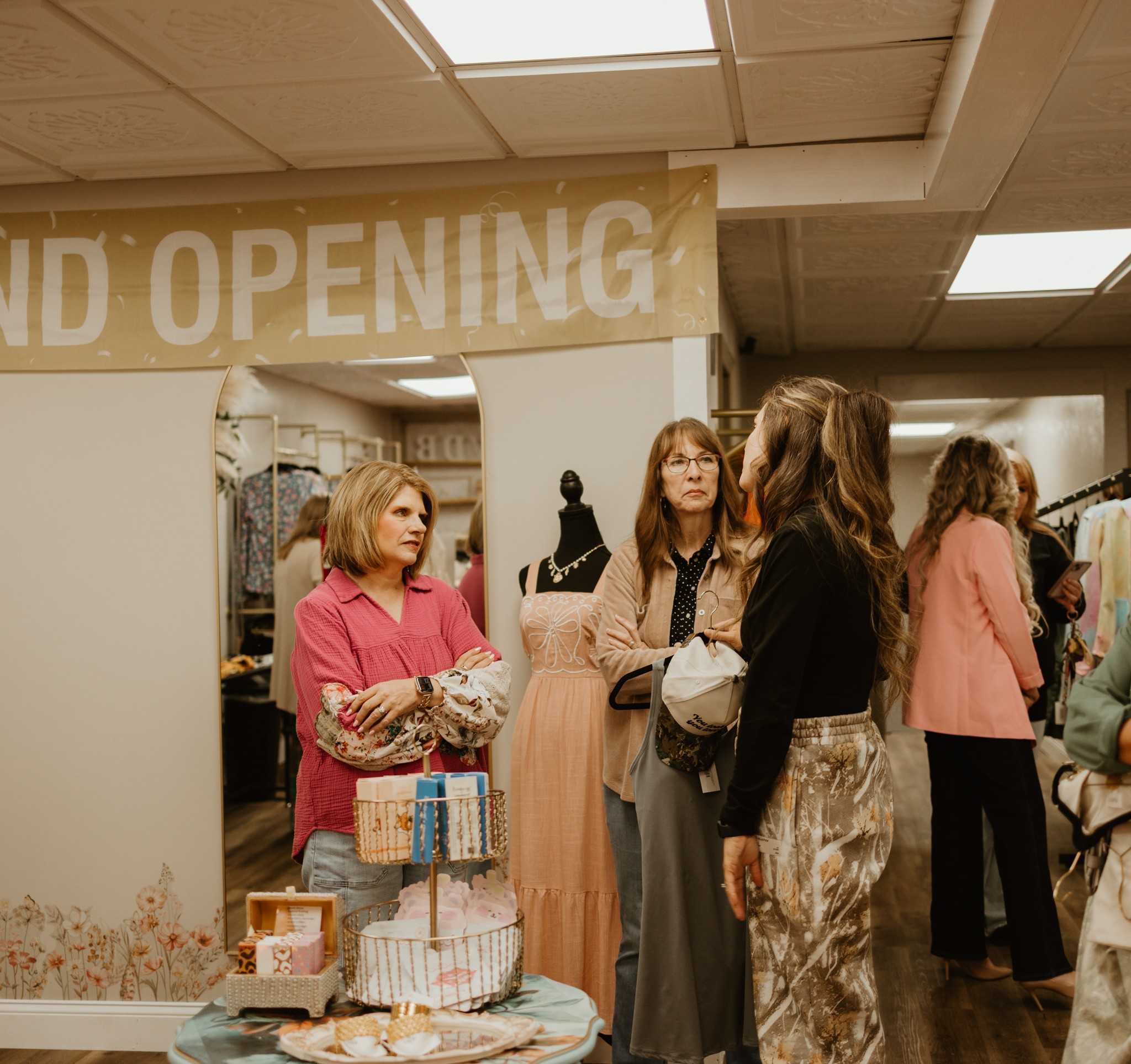 Group of women talking inside a store during grand opening event, with a banner overhead and shopping items on table.