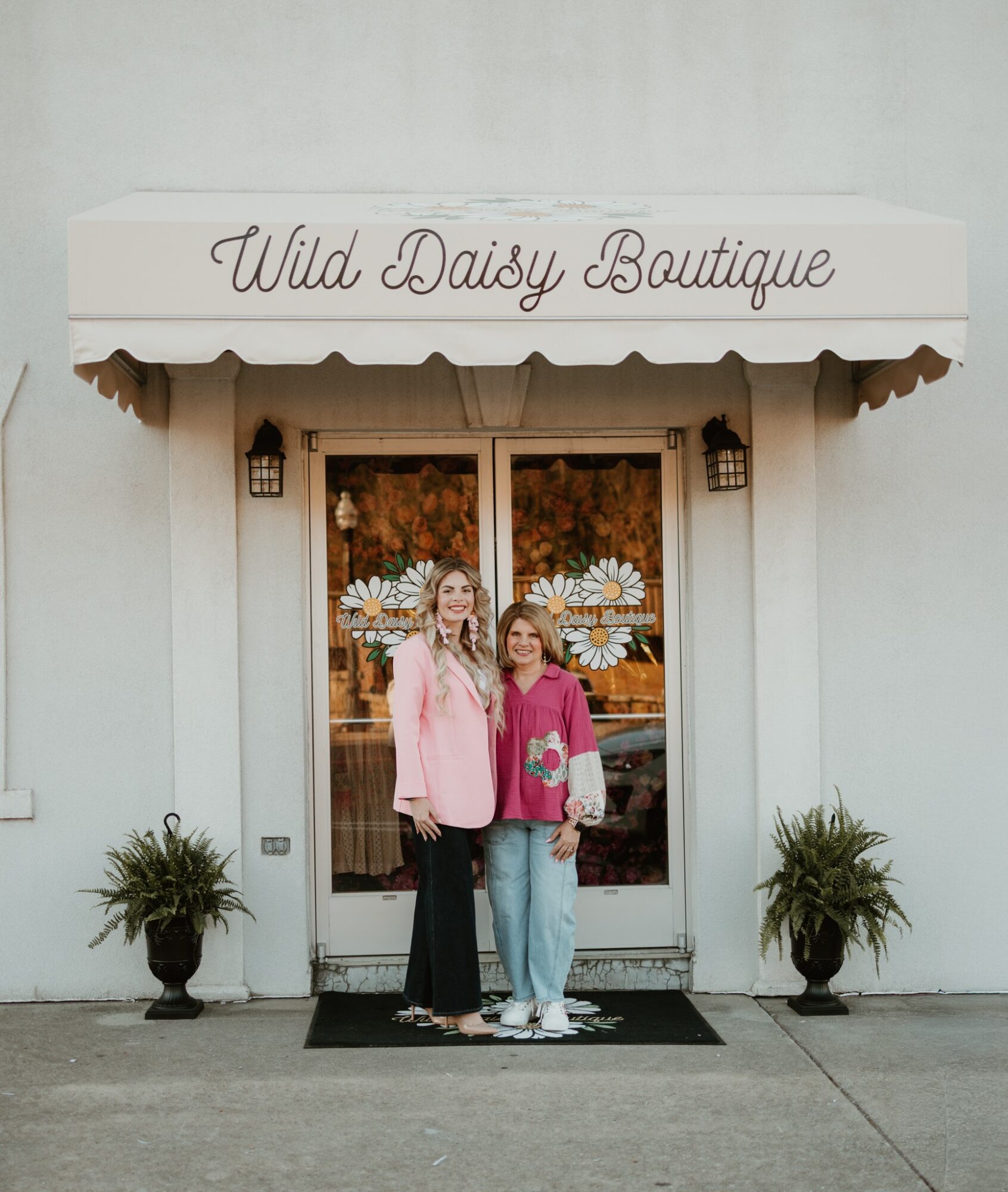 Two women stand in front of a boutique entrance with a sign reading 'Wild Daisy Boutique,' flanked by potted plants.