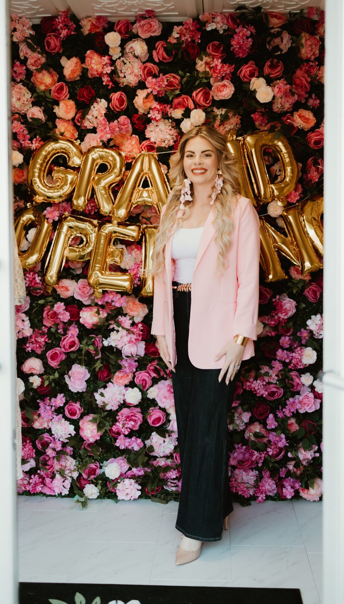 Young woman with long blonde hair in a pink blazer and black pants standing in front of a floral backdrop with gold balloons spelling 'GRAD' and 'PE'.