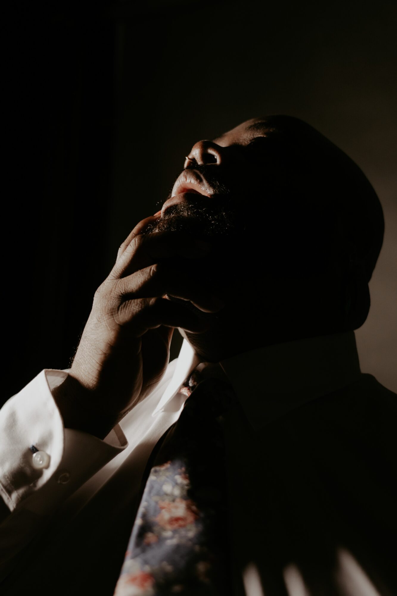 Person with hand on chin, head tilted back, face partially illuminated, dark background, dramatic lighting.