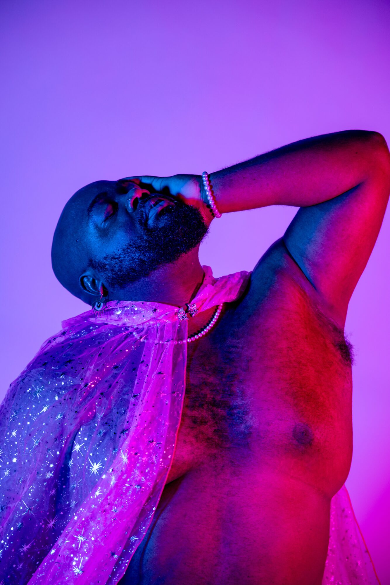 A shirtless person with a glittery cape, head tilted back, hand on forehead, illuminated by colorful lighting.