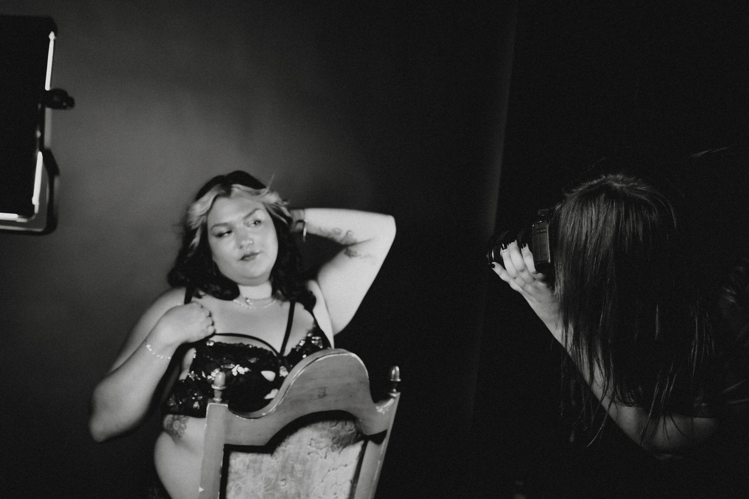 A woman with dark hair is taking a photo of a woman with light hair, who is posing with her hand behind her head.