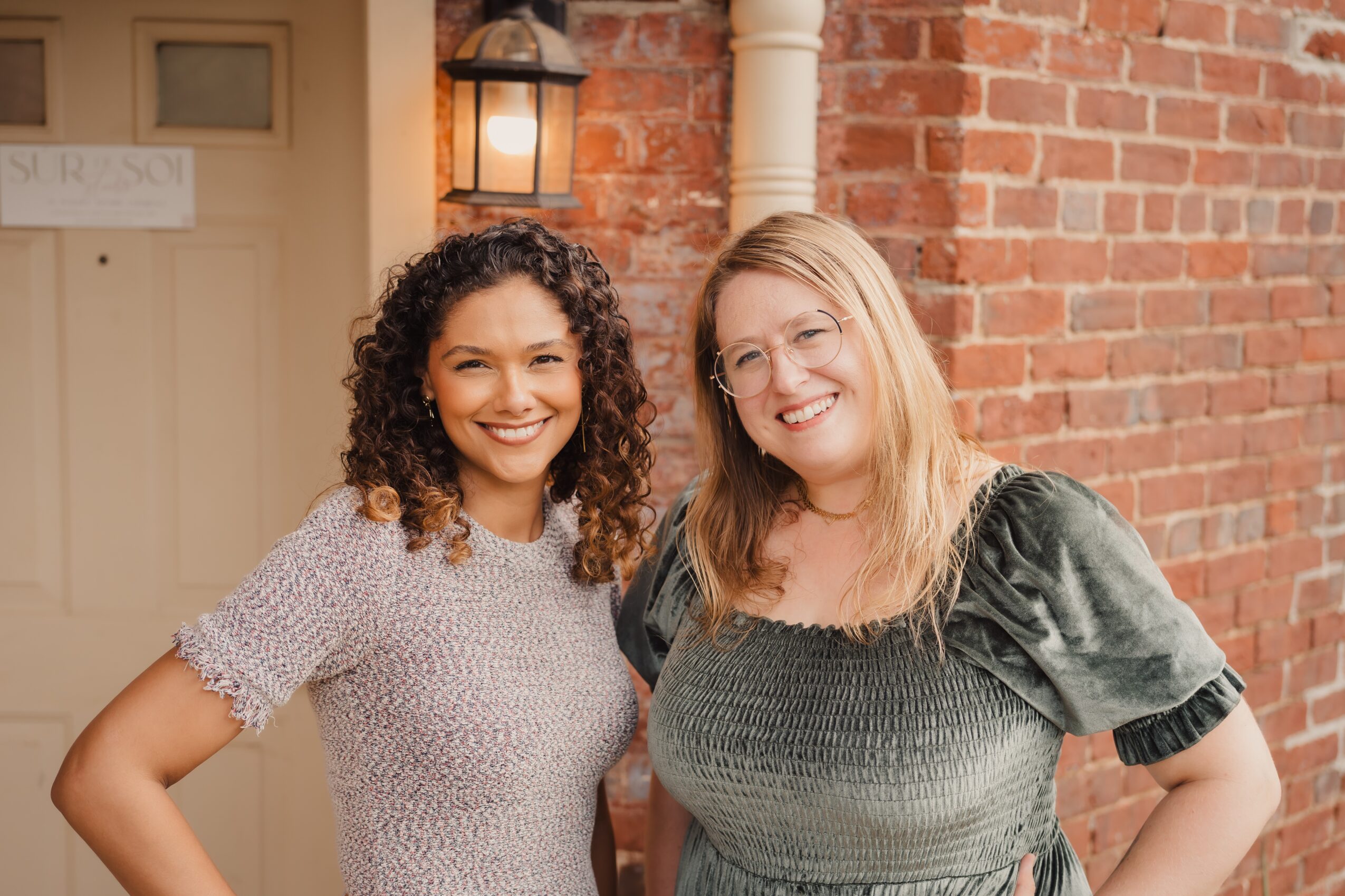Two women smiling and standing close together outdoors against a brick wall, one with curly hair and the other with straight hair.
