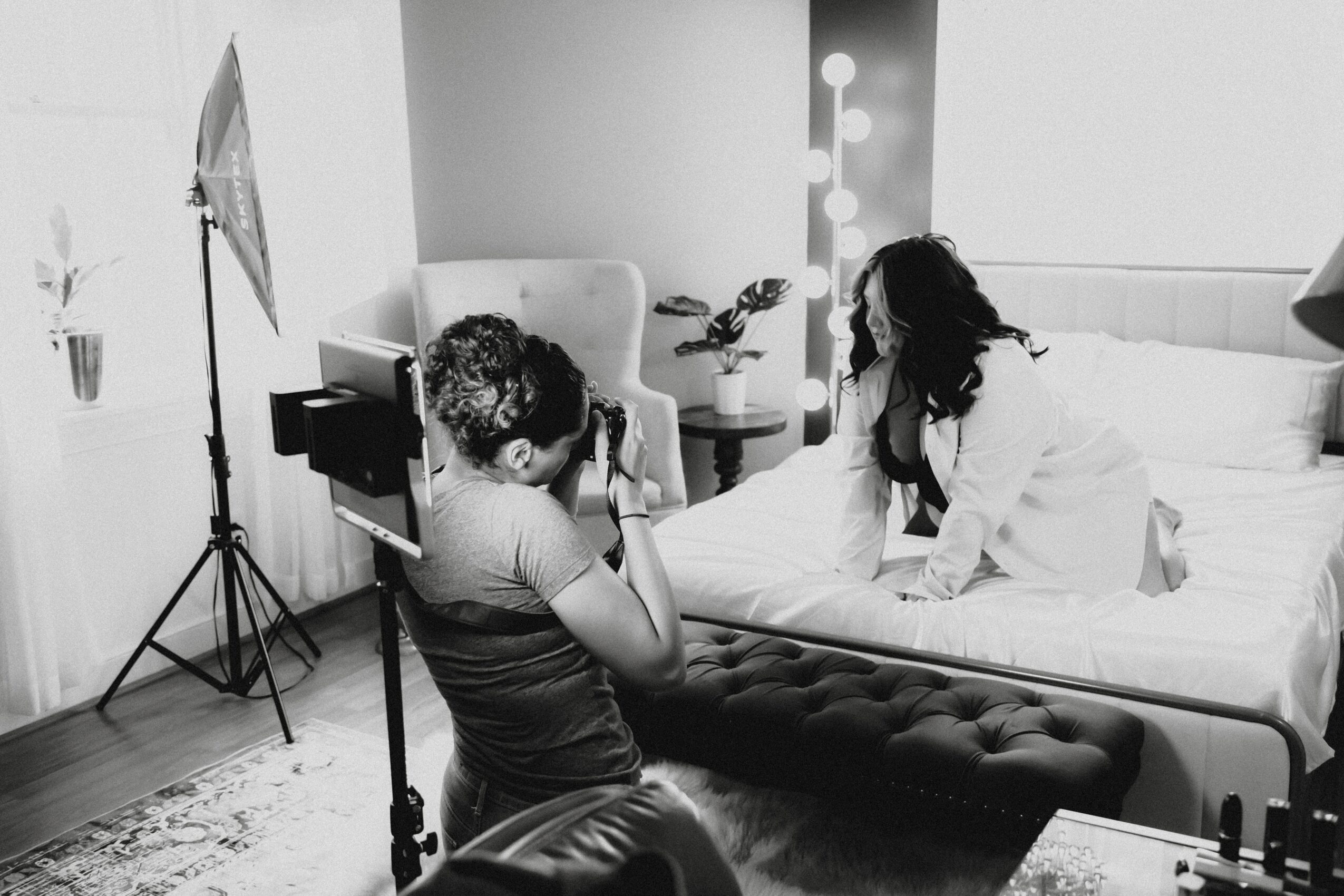 Person taking a photo of a woman on a bed in a bedroom, with a photographer using a camera and lighting equipment, black and white.