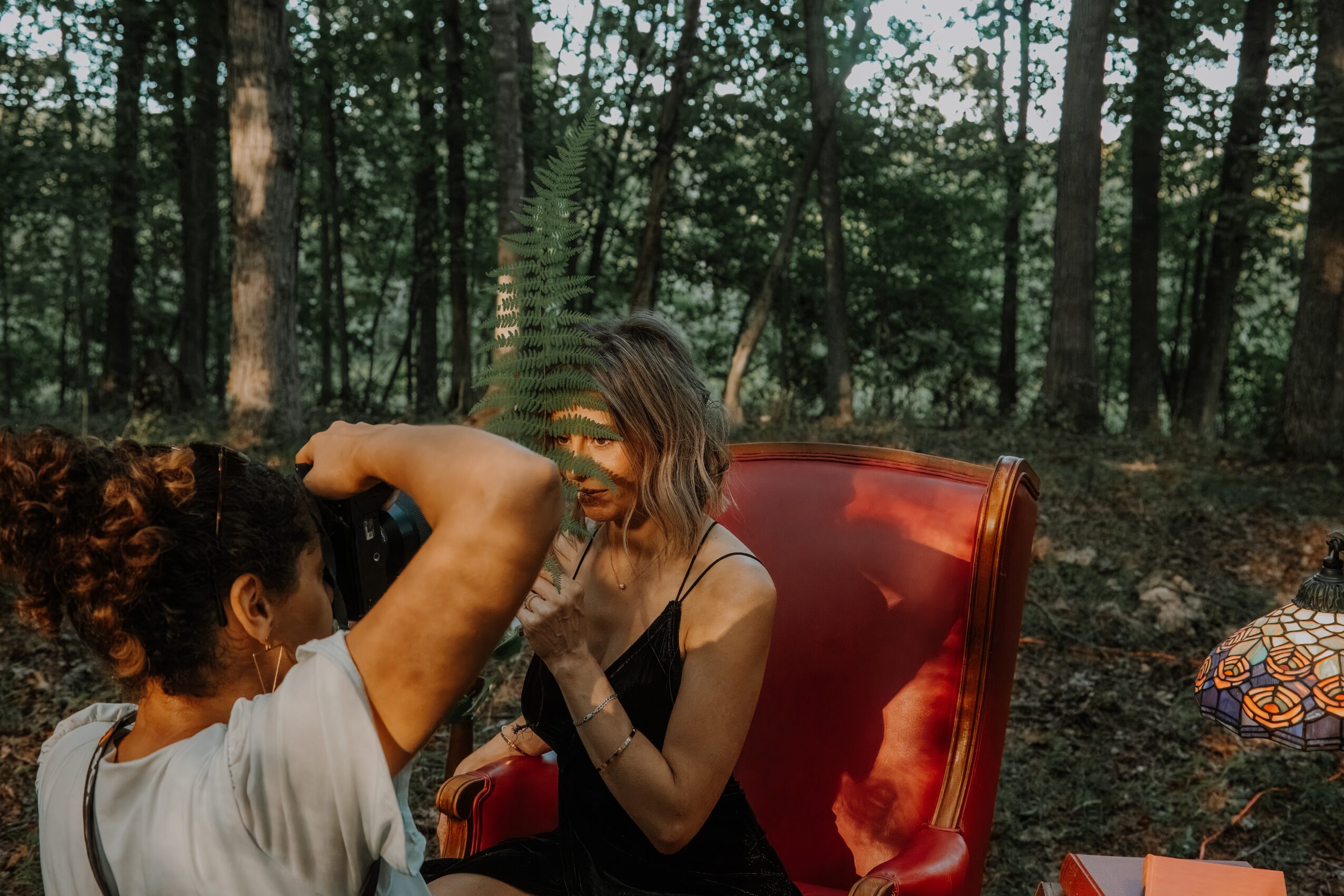 Two women sitting outdoors in a forest, one with curly hair and the other with straight hair, engaging in conversation.