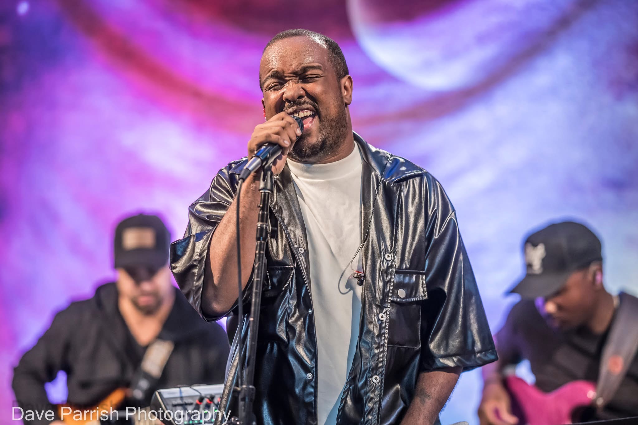 A man singing passionately into a microphone on stage with two musicians in the background, colorful stage lighting.