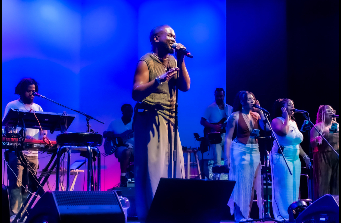 Group of musicians and singers performing on stage with blue lighting, one woman singing into a microphone, others playing instruments.