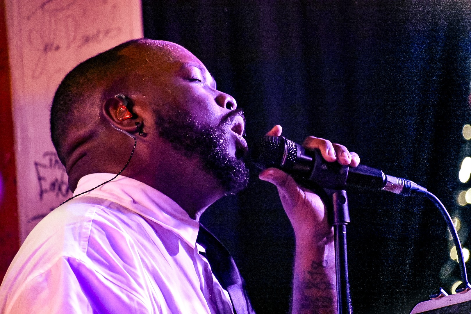 Man singing into a microphone with eyes closed, wearing a white shirt, in a dimly lit setting.