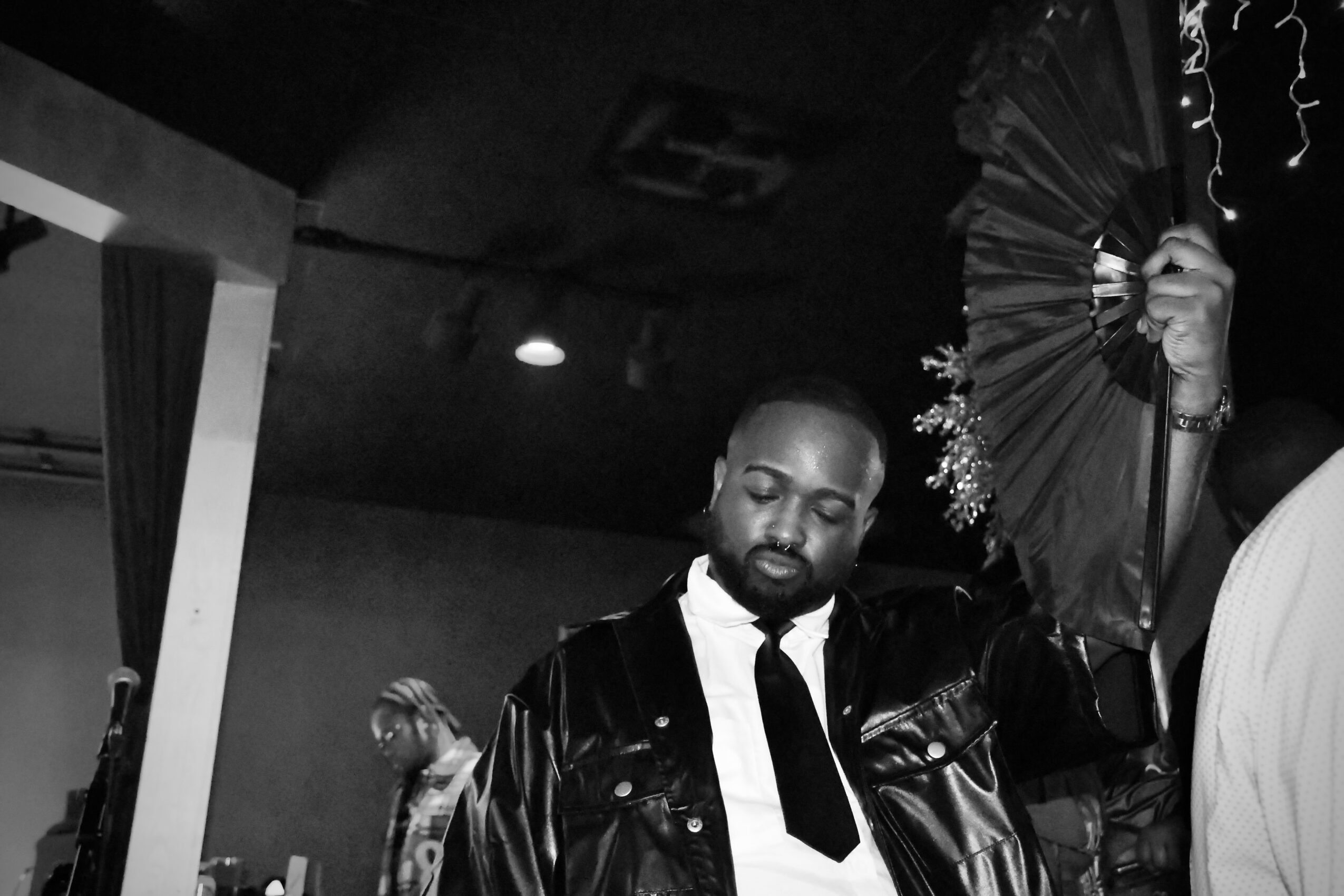 Man with a beard in a leather jacket holding a fan, standing indoors with a dark ceiling and a light fixture, in black and white.