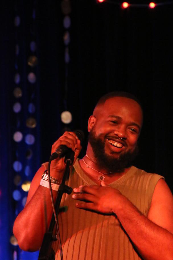 Person smiling, holding a microphone, wearing a sleeveless top, with a dark background and colorful lights.