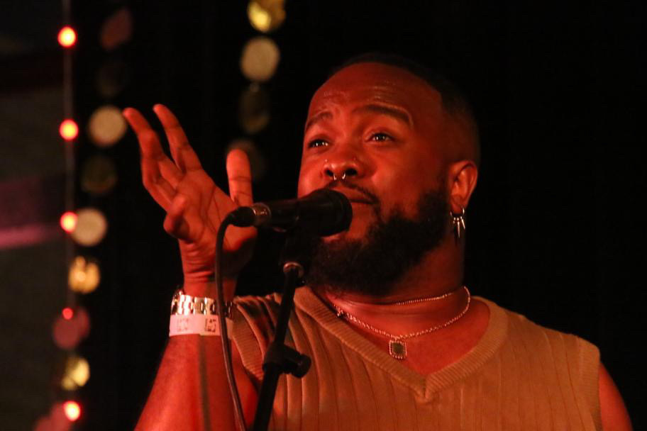 A man with a beard and earrings sings into a microphone on stage, with blurred lights in the background.
