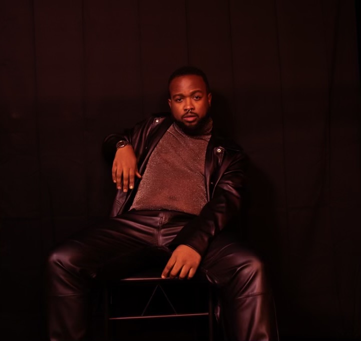 Man sitting on a chair against a dark background, wearing a leather jacket and gray shirt.