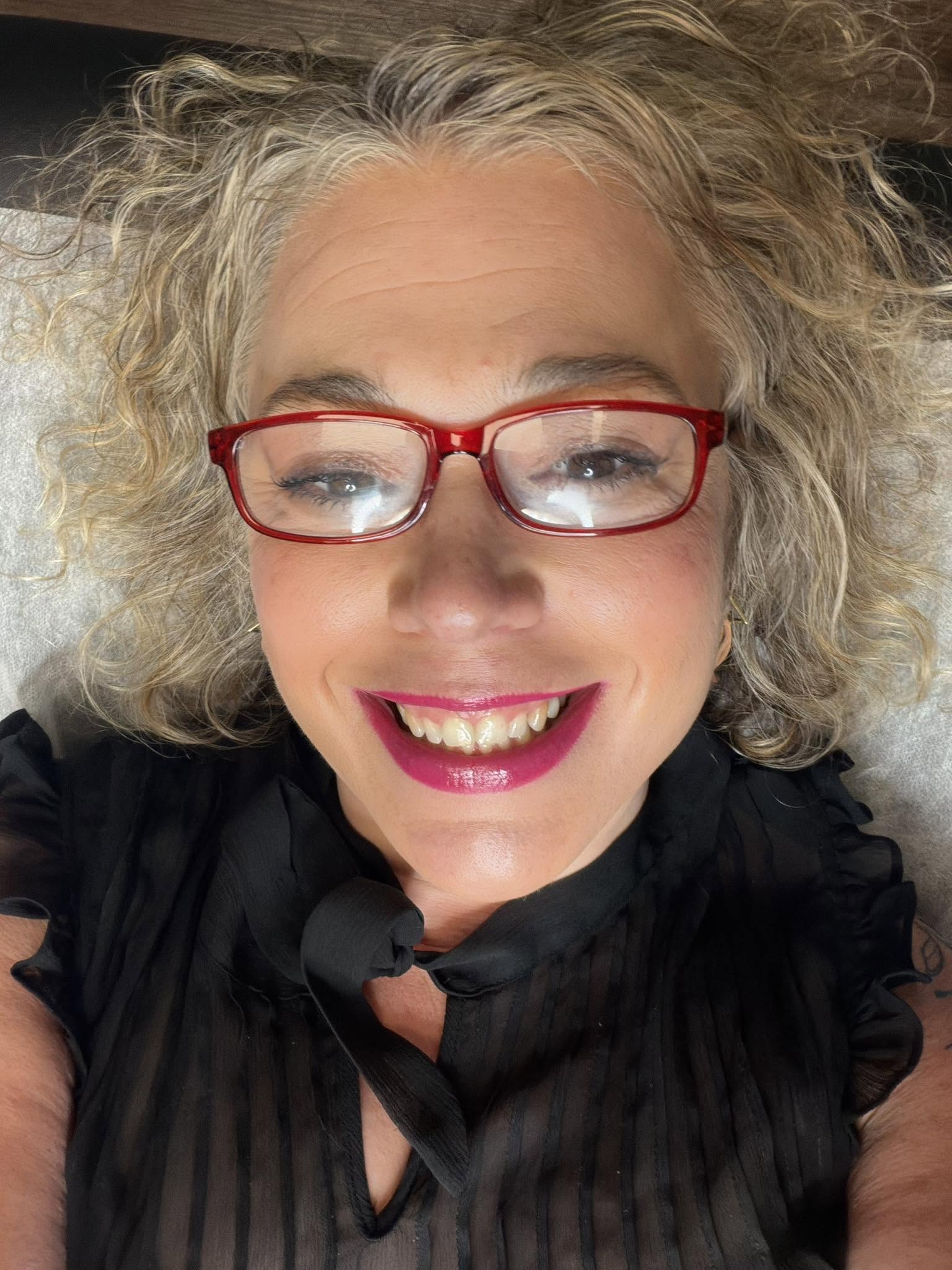 Close-up of a smiling woman with curly gray hair wearing red glasses and a black top.