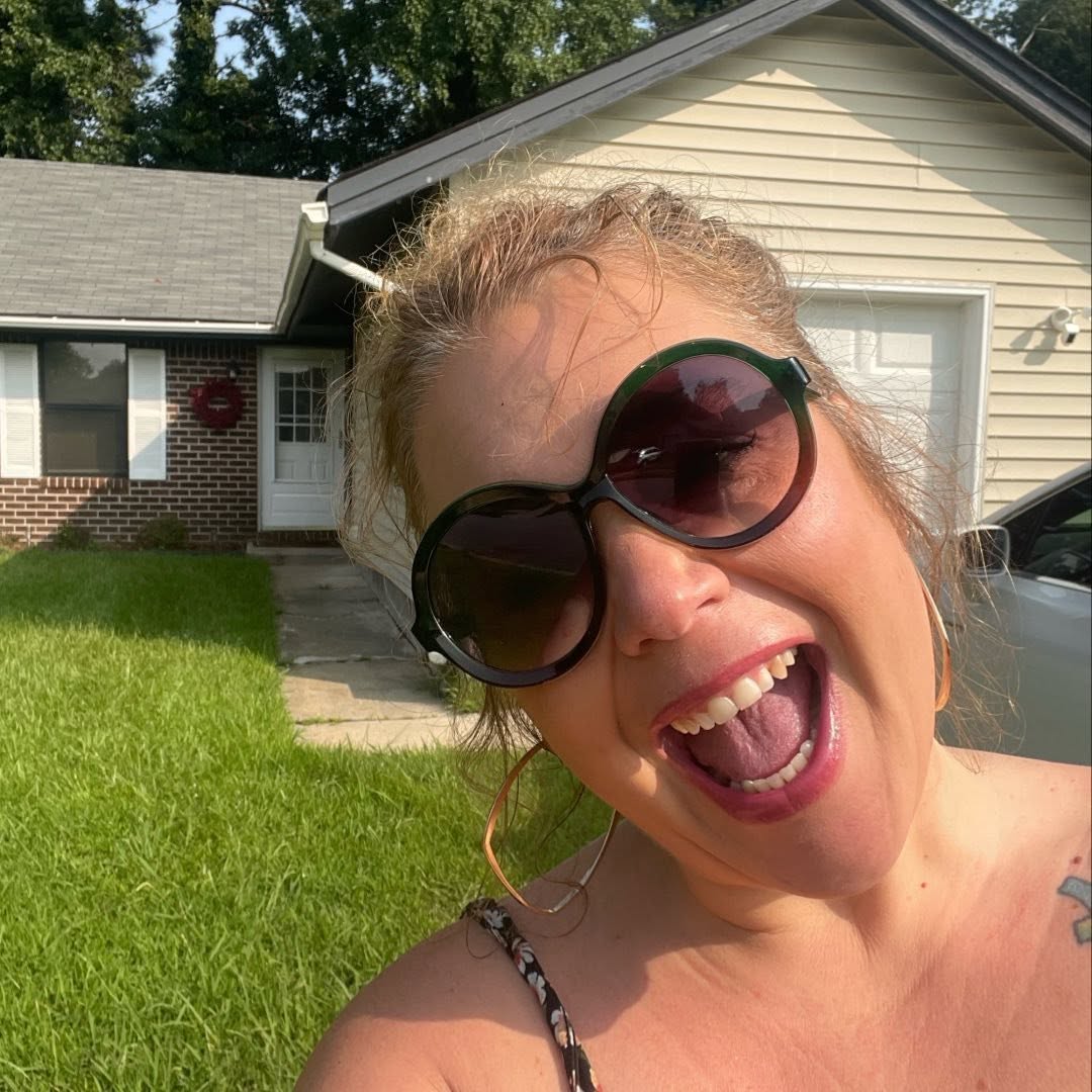 Woman with large sunglasses smiling outdoors in front of a house and green lawn.