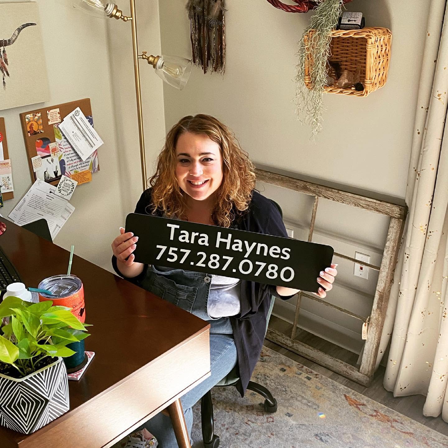 Young woman with curly hair smiling, holding a sign with contact information, sitting at a table in a cozy room.