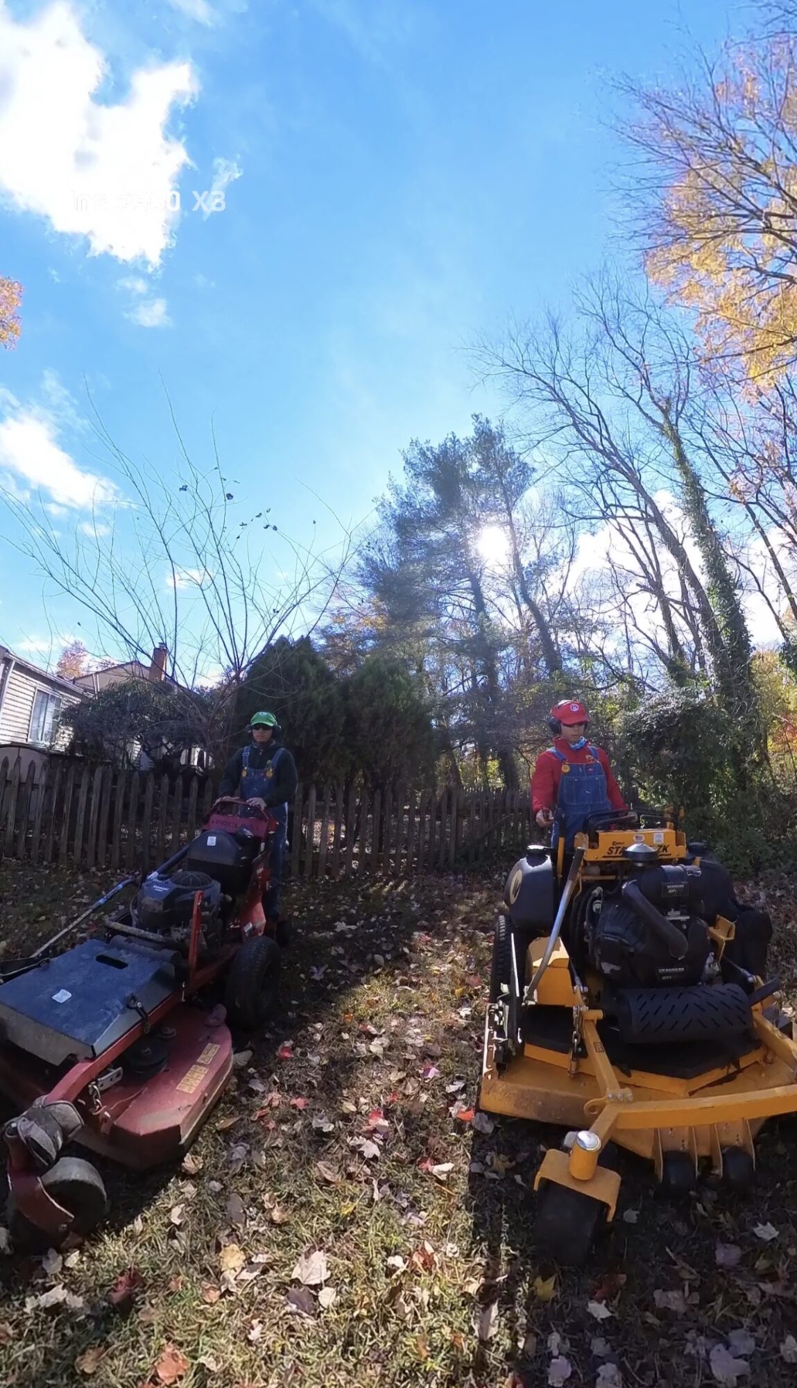 Two people with backpacks and helmets stand outdoors near trees and equipment, under a blue sky with clouds.