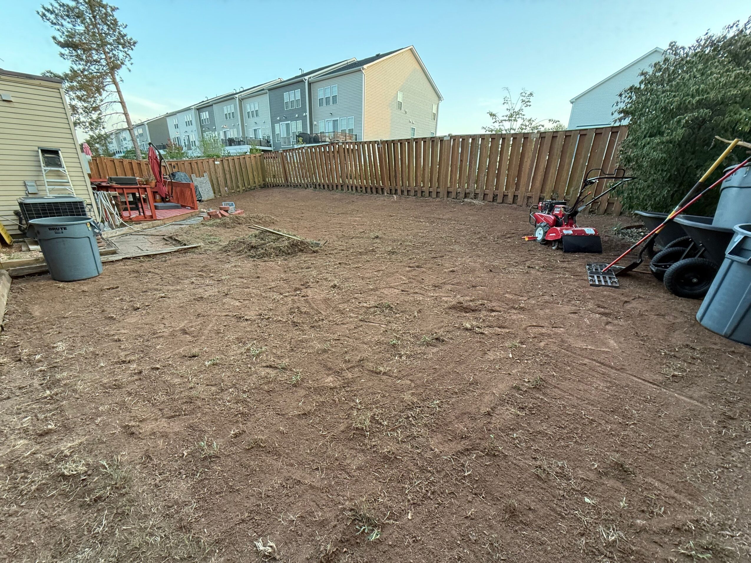 View of a backyard with a dirt ground, wooden fence, and neighboring houses in the background.