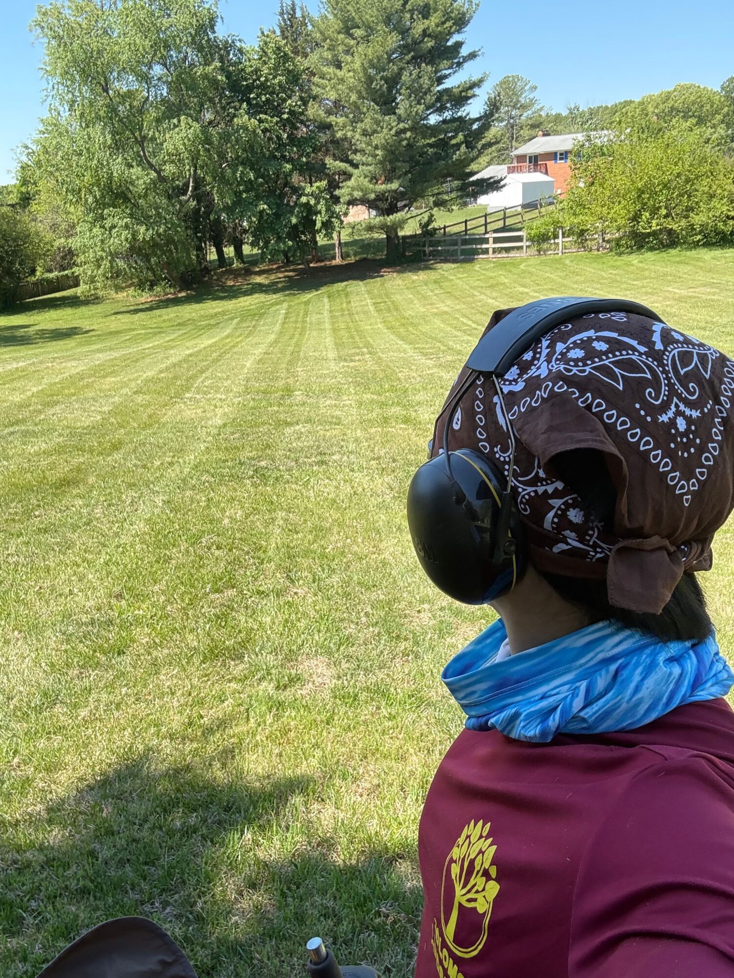 Person wearing a helmet, bandana, and maroon shirt standing on a grassy field with trees and a building in the background.