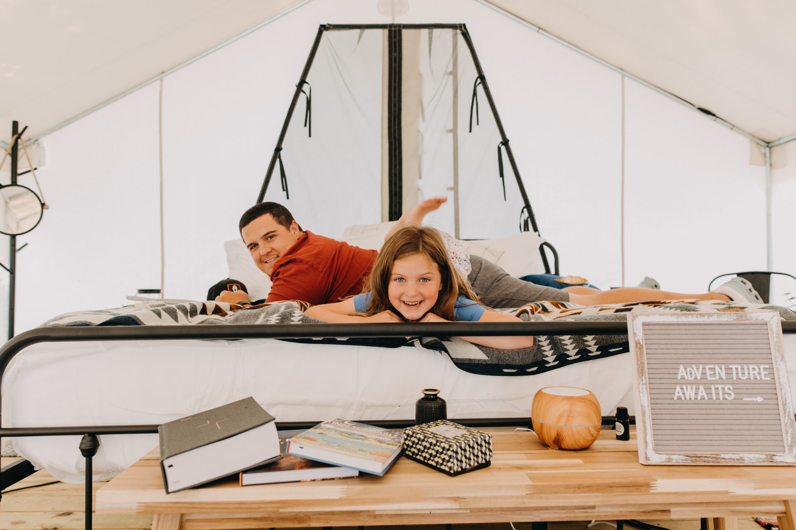 Two children lying on a bed inside a tent, smiling and looking at the camera, with books and a pumpkin on a table nearby.