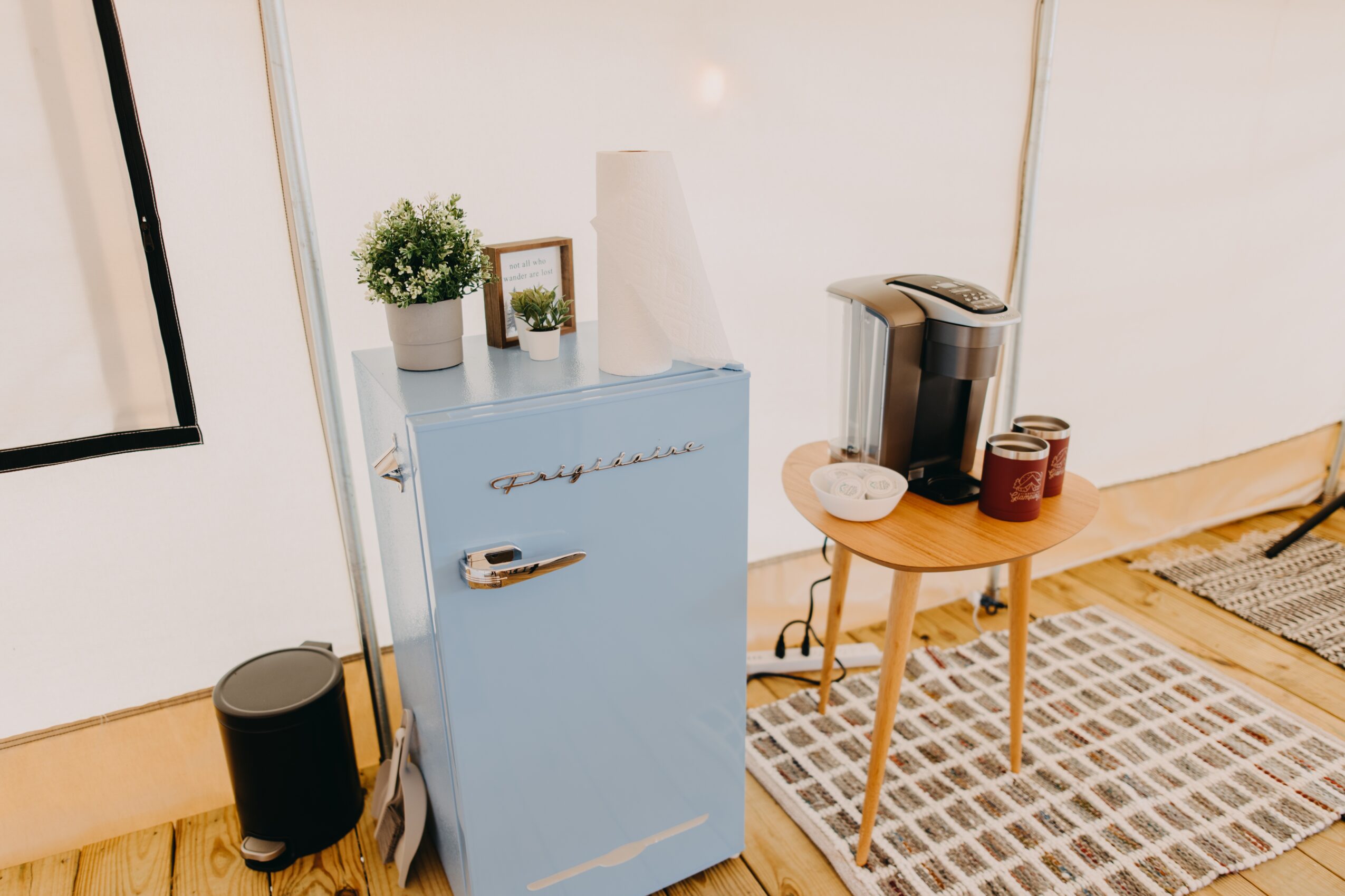 Light blue cabinet with a potted plant, picture frame, and white vase on top, next to a small wooden table with a coffee machine and jars, on a patterned rug.