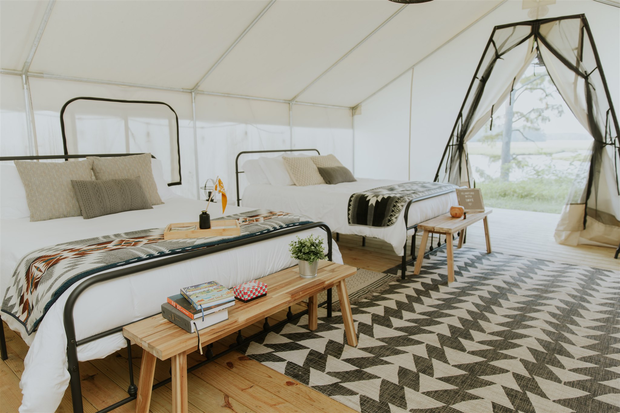 Two beds with pillows and blankets, wooden benches at foot of beds, patterned rug, tent-like window, and natural light.