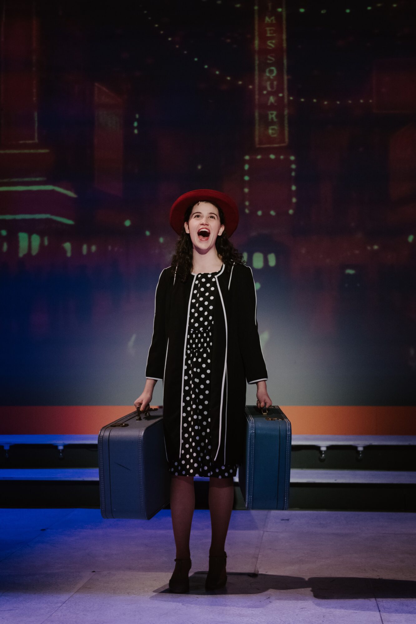 Young woman in a polka dot dress and red hat holding two suitcases, standing on stage with cityscape background.