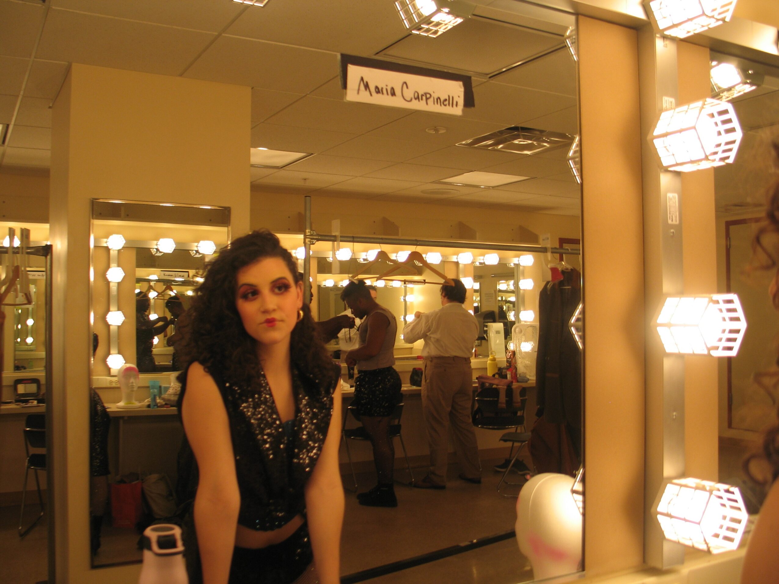Woman with curly hair in front of a mirror with bright lights, backstage dressing room, sign above reads 'Anna Campbell'.