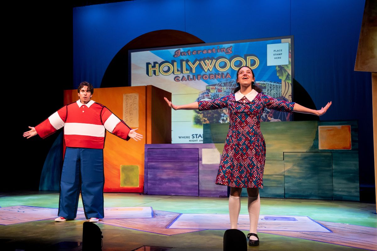 Two performers on stage with colorful blocks and a Hollywood sign backdrop, one with arms outstretched, the other standing with arms open.