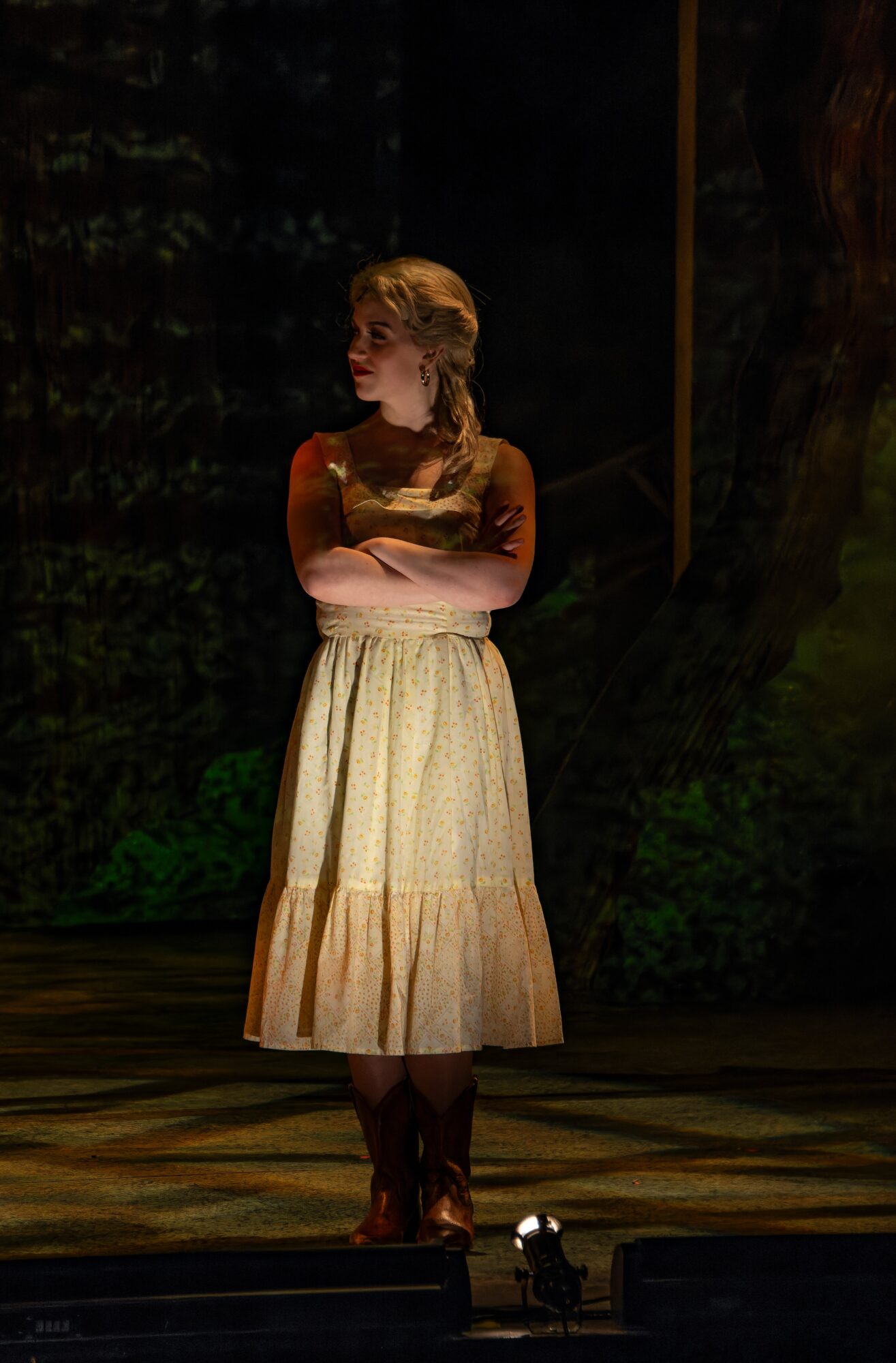 Woman standing on stage with arms crossed, wearing a long skirt and top, in a dark setting with a backdrop.