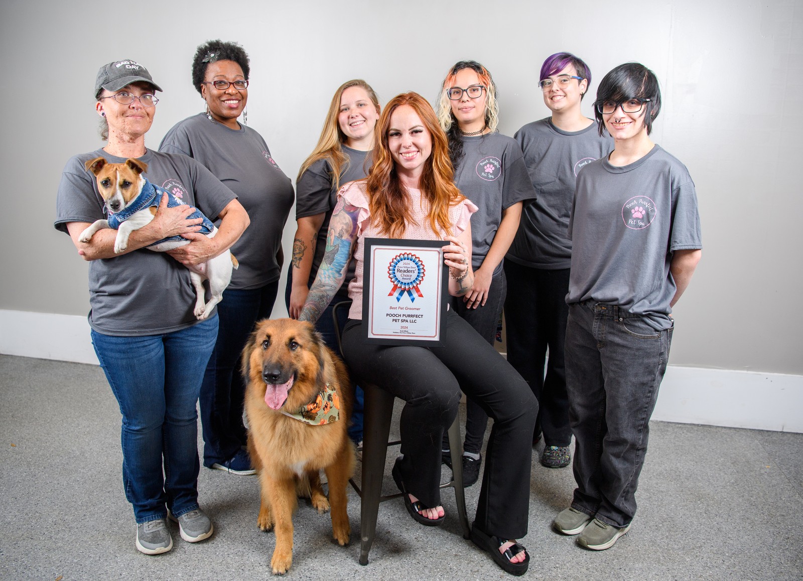 Group of seven people, two dogs, and a framed award, standing against a plain wall, smiling, with one person seated holding the award.
