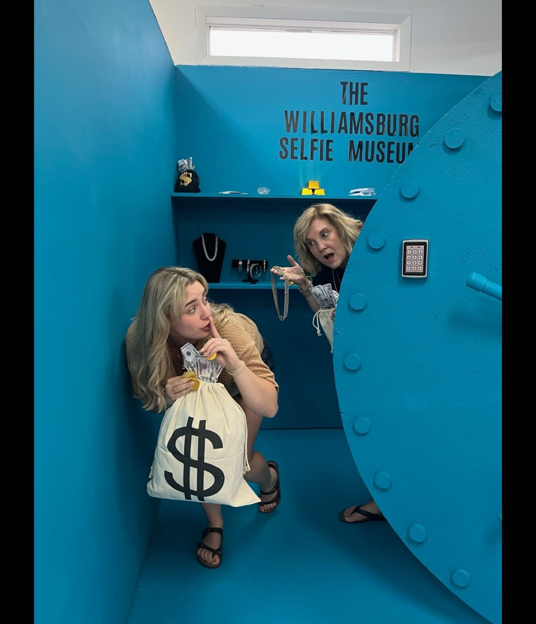 Two women inside a blue room at the Williamsburg Selfie Museum, one holding a money bag prop, the other peeking from behind a large safe door, with a sign on the wall.