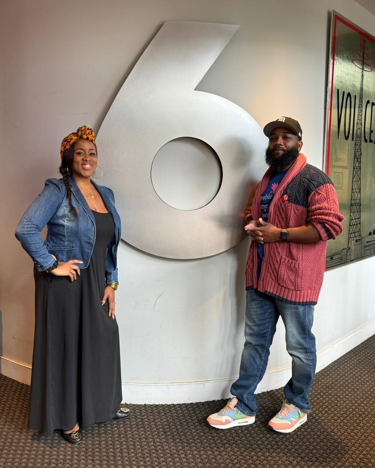 Two people stand beside a large number six sign indoors, smiling and posing for the photo.