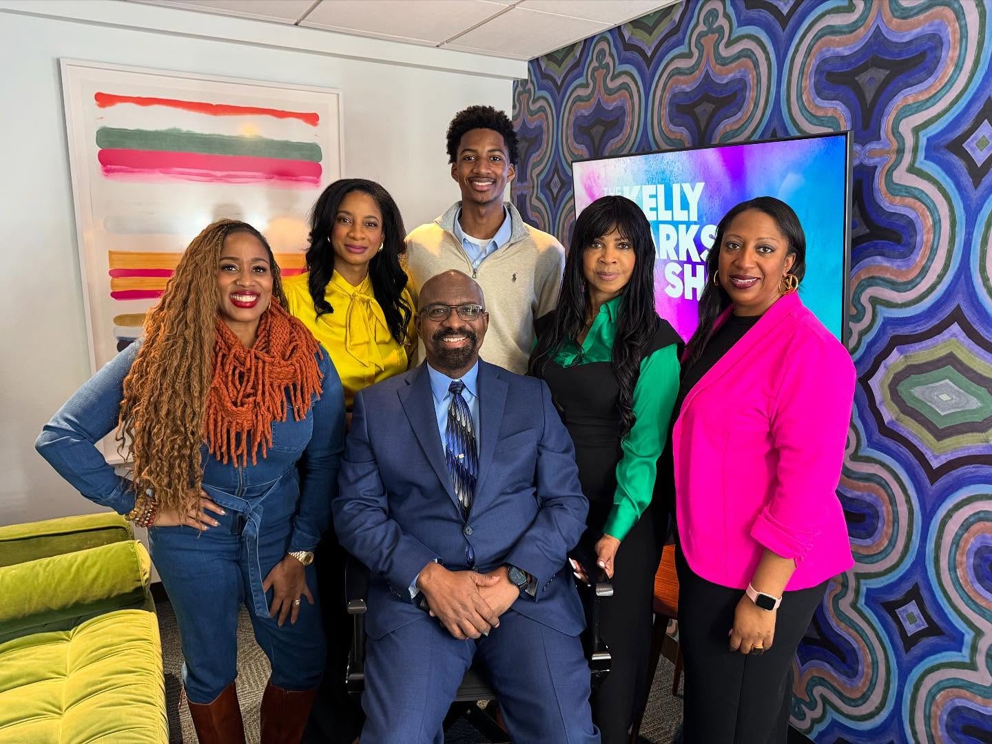 Group of six diverse people smiling in a colorful room with patterned walls and a TV screen, posing for a photo.