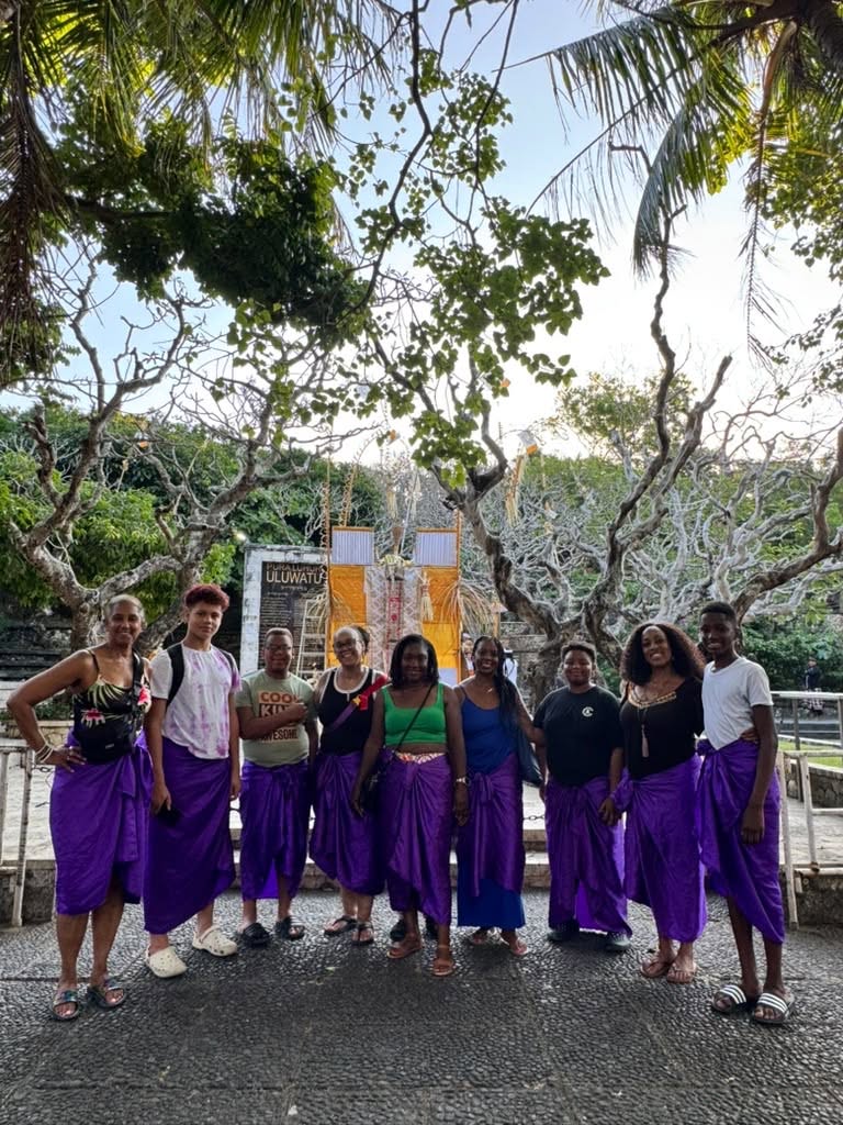 Group of nine women in purple skirts and colorful tops standing outdoors on a sunny day, trees in background.