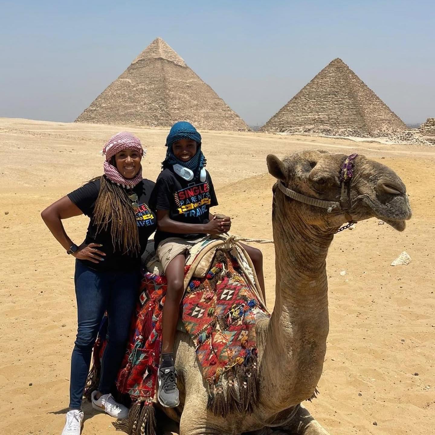 Two women with a camel in front of pyramids in a desert landscape, one sitting on the camel and the other standing beside it.